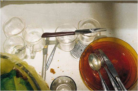 תצלום תקריב ממבט עילי על כלי אוכל שהונחו בתוך כיור מטבח לאחר ארוחה. צלחות זכוכית חומות, כוסות שתייה שקופות, כף, מזלג וסכינים פזורים לצד סלסלת שיירי מזון.